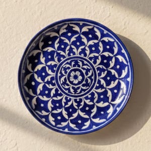 以下が含まれることがあります： 青と白の花柄の丸い装飾皿。 皿はクリーム色の壁に取り付けられています。 ブランド名「AAKARSHAN KALA」が左上に表示されています。