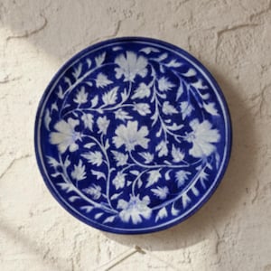 May include: A blue and white decorative plate with a floral design is mounted on a textured wall. Below the plate hangs a macrame wall hanging with a wooden dowel and cream-colored fringe.