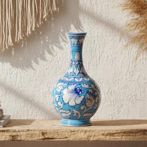 Puede incluir: Un jarrón de cerámica azul y blanco con diseños florales. Se encuentra sobre una estantería de madera junto a una maceta de terracota con hierbas secas, una cesta tejida y libros. El texto "AAKARSHAN KALA" es visible. Un objeto decorativo.
