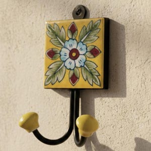 May include: A wall-mounted, double hook with a square, yellow ceramic tile featuring a floral design in blue, green, and red. The hooks are black metal with yellow tips. The brand name "AAKARSHAN KALA" is in the top left corner.