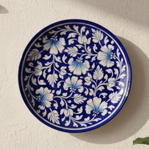 以下が含まれることがあります： 丸い青と白の装飾用プレート。花柄のデザイン。白い壁に取り付けられています。濃い青色の縁があり、白い花と葉で装飾されています。「AAKARSHAN KALA」のブランド名が左上にあります。