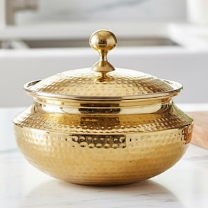 May include: A gold-coloured, hammered-metal lidded bowl with a round finial. The bowl is on a white countertop. The background includes a cutting board, a small potted plant, and a kitchen sink.
