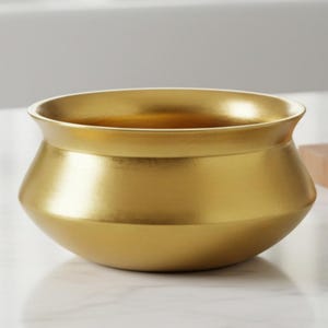 May include: A gold-colored, decorative bowl with a wide rim and a rounded body. The bowl is sitting on a white countertop in a kitchen setting. The brand name "AAKARSHAN KALA" is visible in the top left corner.