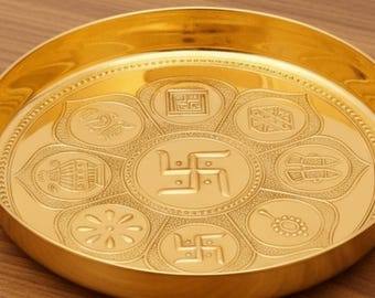 May include: A gold-colored, round decorative plate with embossed religious symbols. The plate has a raised rim and is set on a wooden surface. The text "AAKARSHAN KALA" is in the upper left corner.