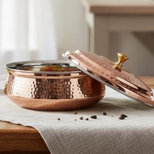 Puede incluir: Un cuenco de servir de color cobre con una textura martillada y una tapa a juego. La tapa está ligeramente entreabierta, revelando el contenido del cuenco. El cuenco está sobre un paño de lino de color neutro sobre una mesa de madera. El texto "AAKARSHAN KALA" está arriba a la izquierda.