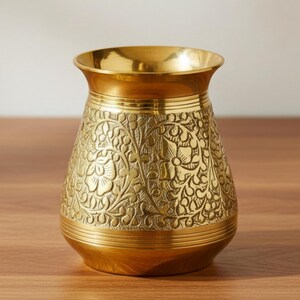May include: A gold-colored metal vase with intricate floral patterns etched into its surface. The vase has a wide, rounded base and a flared rim. The text "AAKARSHAN KALA" is in the upper left corner.