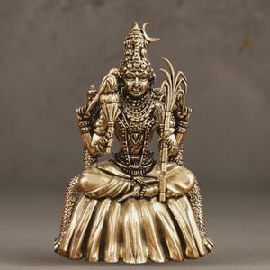 May include: A gold-colored statue of a seated deity, adorned with intricate jewelry and a crescent moon atop the head. The figure holds objects in multiple hands and is seated on a detailed, flowing garment. The text "AAKARSHAN KALA" is in the upper left corner.