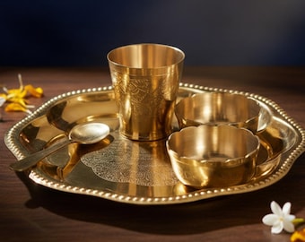 May include: A gold-colored brass dining set with a decorative tray, a cup, two small bowls, and a spoon. The tray has a beaded edge. The set is displayed on a dark wooden surface with flowers and the text "AAKARSHAN KALA".