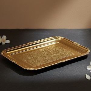 Puede incluir: Una bandeja rectangular dorada con detalles ornamentados y un patrón floral. La bandeja tiene un borde decorativo y está colocada sobre una superficie oscura. El texto "AAKARSHAN KALA" está en la esquina superior izquierda.
