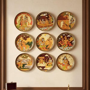 May include: Nine decorative round plates with colourful illustrations of religious figures and scenes. The plates are arranged in a grid on a beige wall. The plates have a gold-coloured rim. A dark wooden cabinet is below the plates.
