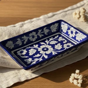 Pode incluir: Uma travessa retangular de cerâmica com borda azul escuro e padrões florais brancos. A travessa está colocada sobre um tecido bege, com uma superfície de madeira ao fundo. A marca "Aakarshan Kala" é visível no canto superior esquerdo.