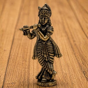 May include: A metallic gold-toned figurine of a deity playing a flute, featuring intricate carvings on the clothing and crown. The figure stands in a crossed-leg posture. The text AAKARSHAN KALA is visible in the upper left corner of the image.