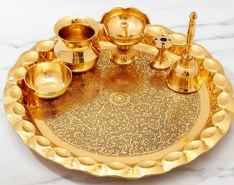 Op de afbeelding: Een goudkleurige puja thali set met diverse componenten, waaronder een kleine kom, een bel, een lamp en een kleine pot. De thali heeft een decoratief bloemmotief en een geschulpte rand. De tekst "AAKARSHAN KALA" is linksboven zichtbaar.