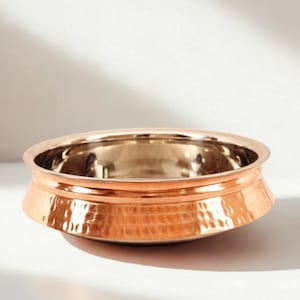 May include: A copper-colored bowl with a hammered texture on the exterior. The bowl has a wide, shallow shape with a polished interior. The brand name "AAKARSHAN KALA" is visible in the top left corner. The bowl is likely for serving food.