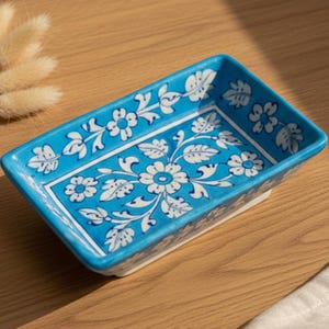 Puede incluir: Un plato rectangular de cerámica pintado a mano, con un fondo azul vibrante y diseños florales blancos. El plato tiene un borde elevado y se muestra sobre una superficie de madera. El texto "AAKARSHAN KALA" es visible en la esquina superior izquierda.