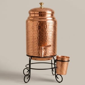 May include: A hammered copper water dispenser with a lid and a spigot, resting on a black metal stand that includes a matching hammered copper cup holder. The text AAKARSHAN KALA is visible in the upper left corner of the image.