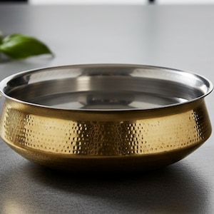 Puede incluir: Un cuenco redondo de dos tonos con exterior dorado martillado e interior plateado. El cuenco tiene un diseño ancho y poco profundo y un borde ligeramente ensanchado. La marca "AAKARSHAN KALA" es visible en la esquina superior izquierda.