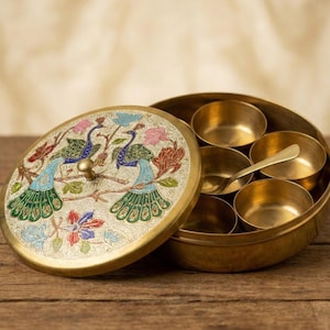 May include: A brass spice box with a lid featuring a peacock design in blue, green, and pink. The box contains small brass bowls and a spoon. The text "AAKARSHAN KALA" is visible on the top left corner.