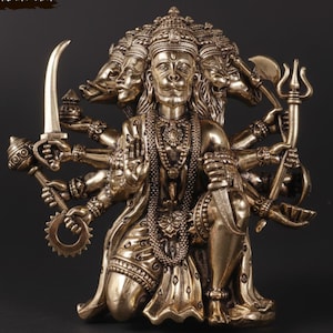 Puede incluir: Estatua de color bronce de una deidad con múltiples brazos y rostros, sosteniendo varias armas y símbolos. La estatua está arrodillada y adornada con intrincadas joyas. El texto "AAKARSHAN KALA" es visible en la esquina superior izquierda. Fondo oscuro.