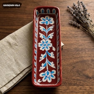 May include: A rectangular, maroon ceramic dish with a floral design in white and blue. The dish has a raised rim and is decorated with stylized flowers and leaves. The text "AAKARSHAN KALA" is in the upper left corner.