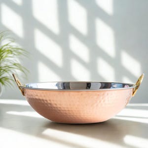 May include: A copper-coloured, hammered metal bowl with gold-coloured handles. The bowl has a shiny, silver interior. The image is of a kitchenware item, likely for cooking or serving food. The brand name "Aakarshan Kala" is visible in the top left corner.