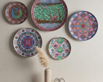 May include: A collection of five decorative plates with colorful, intricate designs. The plates feature various patterns, including birds, trees, and geometric shapes. The plates are displayed on a neutral-colored wall, with a vase and decorative object on a wooden shelf.