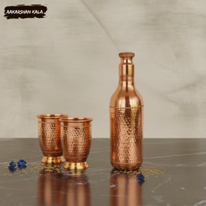 May include: A copper water bottle and two matching cups with a hammered texture. The bottle has a long neck and a decorative stopper. The cups have a flared shape and a gold-coloured base. The brand name "AAKARSHAN KALA" is visible.
