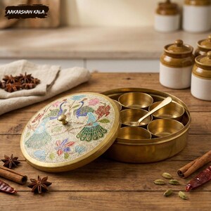 May include: A brass spice box with a decorative lid featuring peacock designs in blue, green, and pink. The box contains small brass bowls and a spoon. The text "AAKARSHAN KALA" is visible in the top left corner. The box is on a wooden surface.