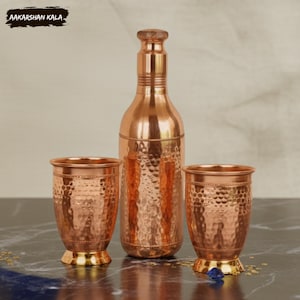Może przedstawiać: Miedziana butelka na wodę z dwoma pasującymi miedzianymi szklankami. Butelka i szklanki mają młotkowaną fakturę i złotą podstawę. Nazwa marki "AAKARSHAN KALA" znajduje się w lewym górnym rogu.