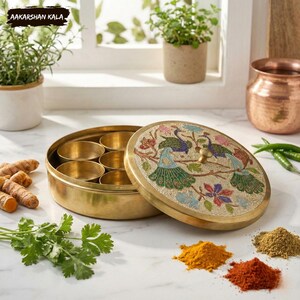 May include: A brass spice box with a lid featuring a peacock design. The box is open, revealing several small compartments. Turmeric, coriander, and other spices are arranged around the box. The text "AAKARSHAN KALA" is in the top left corner.