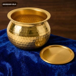 May include: A golden brass pot with a hammered texture and a matching lid. The pot has a wide rim and a rounded body. The lid is a flat, circular disc. The pot and lid are on a blue velvet surface. The text "AAKARSHAN KALA" is in the top left corner.