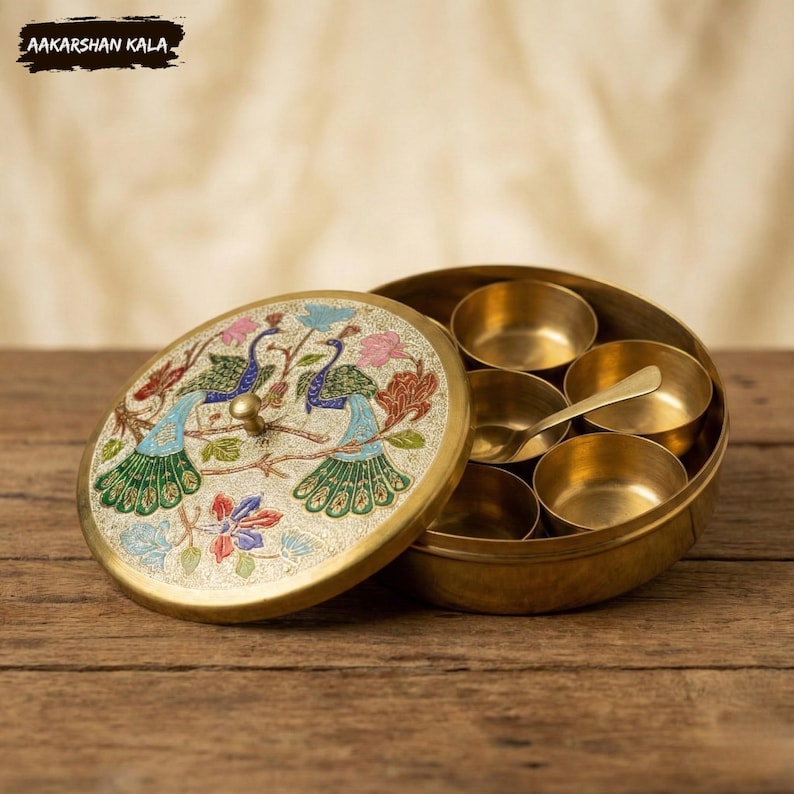May include: A brass spice box with a lid featuring a peacock design in blue, green, and pink. The box contains small brass bowls and a spoon. The text "AAKARSHAN KALA" is visible on the top left corner.