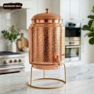 May include: A hammered copper beverage dispenser with a matching lid and a metal spigot, resting on a circular gold-toned wire stand. The text AAKARSHAN KALA is visible in the upper left corner of the frame. The dispenser has a cylindrical body.