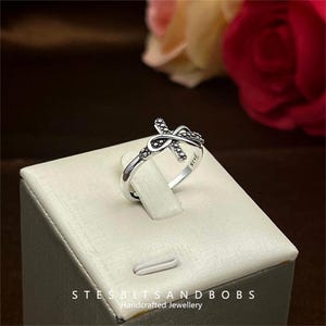 Può includere: Anello d'argento con un design a forma di croce e simbolo di infinito. L'anello è su un espositore bianco con il testo "STESBITSANDBOBS Gioielli artigianali".