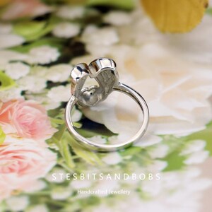 May include: A silver ring with a heart-shaped design. The ring features two heart-shaped elements joined together, with a delicate band. The background is a soft floral pattern. The text "STESBITSANDBOBS Handcrafted Jewellery" is visible.