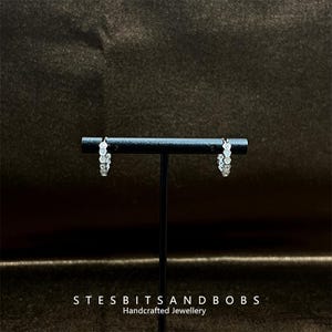 May include: A pair of silver hoop earrings with a row of clear cubic zirconia stones. The earrings are displayed on a black stand against a dark background.