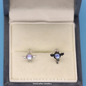 Peut inclure: Deux bijoux de corps argent&eacute;s et noirs avec des pierres iris&eacute;es. La pi&egrave;ce de gauche a des ailes d'ange et la pi&egrave;ce de droite a des ailes de chauve-souris. Les bijoux sont pr&eacute;sent&eacute;s dans un coffret en velours blanc avec les mots "STESBITS AND BOB'S Bijoux artisanaux".