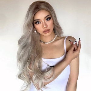 May include: A woman with long, wavy, ash blonde hair and full makeup, including dramatic eye makeup and glossy lips. She wears a white tank top and a pearl and silver chain necklace. Her nails are long and painted with a black and white design.