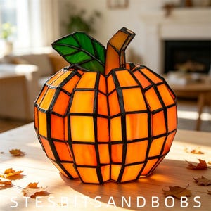 May include: A stained glass pumpkin lamp featuring orange glass panels joined by dark metal solder lines. The pumpkin has a green glass leaf and an orange glass stem. The text STESBITSANDBOBS is visible at the bottom of the image.