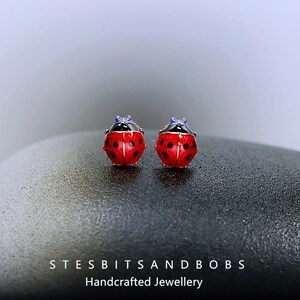 May include: A pair of ladybug stud earrings. Each earring has a red body with black spots, a black head, and blue accents. The earrings are handcrafted jewellery. The text "STESBITSANDBOBS" and "Handcrafted Jewellery" are at the bottom.