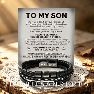 Könnte beinhalten: Ein schwarzes geflochtenes Lederarmband mit einem Metallkreuz-Anhänger und einem Magnetverschluss, der mit J.B. und einem Herzsymbol graviert ist. Das Armband liegt vor einer weißen Karte mit dem Text TO MY SON und einer Botschaft.