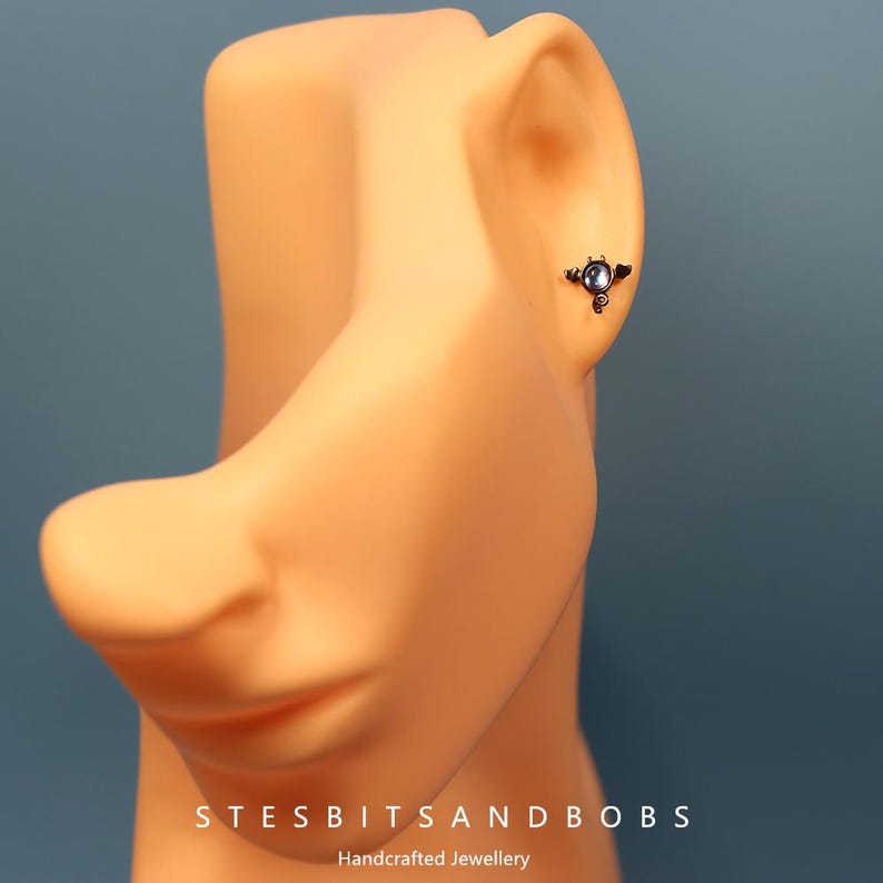 Peut inclure: Gros plan sur une oreille avec un piercing d'oreille en argent et bleu avec une petite pierre pr&eacute;cieuse. Le piercing est un design d&eacute;licat en trois parties avec une petite pierre pr&eacute;cieuse ronde au centre.
