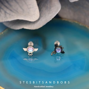 Könnte beinhalten: Zwei silberne und schwarze Körperschmuckstücke mit schillernden blauen Steinen. Ein Stück zeigt ein kleines Engelsflügel-Design und das andere ein kleines Skorpion-Design.