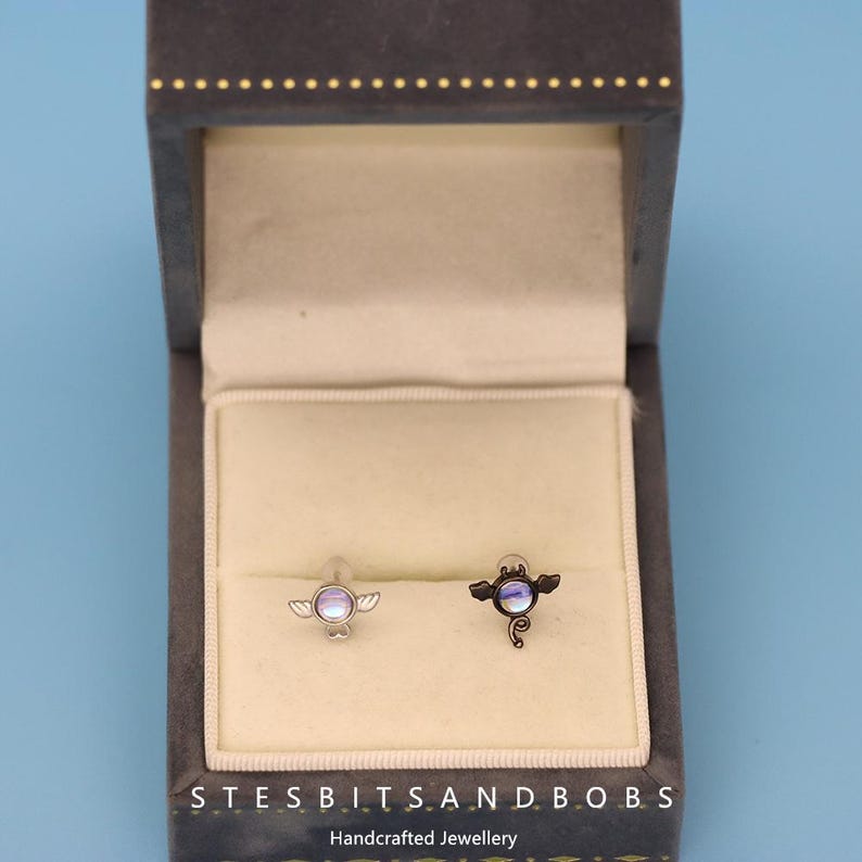 Peut inclure: Une paire de boucles d'oreilles en argent dans un petit coffret en velours. Une boucle d'oreille repr&eacute;sente un ange en argent avec une pierre bleu iris&eacute;e, et l'autre boucle d'oreille repr&eacute;sente un diable en argent avec une pierre bleu iris&eacute;e.