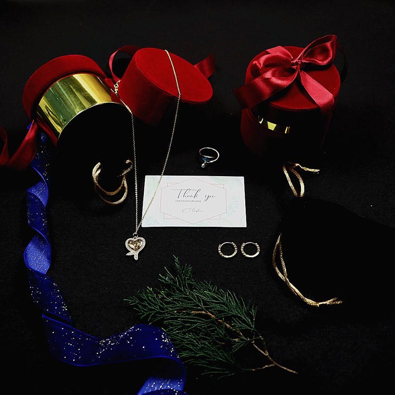 May include: An assortment of jewelry and gift boxes on a black surface. The jewelry includes a heart pendant necklace, hoop earrings, a ring, and two pairs of small hoop earrings. Red velvet gift boxes with gold accents and a blue ribbon are also present.