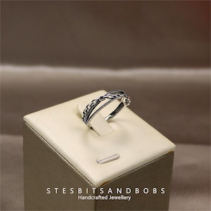 Puede incluir: Un anillo de plata con un diseño de triple cadena. El anillo se muestra en un expositor blanco con el texto "STESBITSANDBOBS Joyería artesanal".