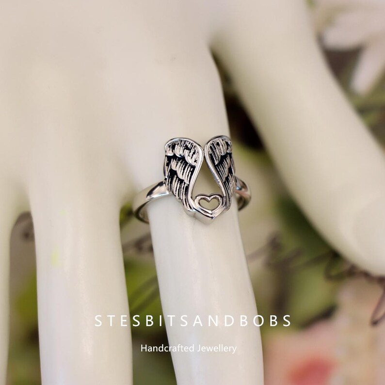 May include: A silver-colored ring featuring an open design of angel wings with a heart at the base. The ring is worn on a finger. The text "STESBITSANDBOBS" and "Handcrafted Jewellery" are visible.