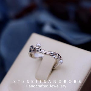 May include: A silver ring with a fluid, organic form, featuring three small pearls. The ring's surface is reflective, and the text "STESBITSANDBOBS Handcrafted Jewellery" is displayed at the base of the image.