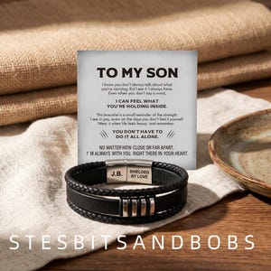 May include: A black leather bracelet with a braided texture and a stainless steel clasp. The clasp is engraved with the initials J.B. and the text SHIELDED BY LOVE. It is displayed in front of a card with the printed message TO MY SON.
