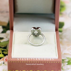 May include: A silver ring with a heart-shaped design, set inside a pink jewelry box. The ring's heart shape is composed of two larger heart shapes and a smaller heart in the center. The jewelry box has the text "STESBITSANDBOBS Handcrafted Jewellery".